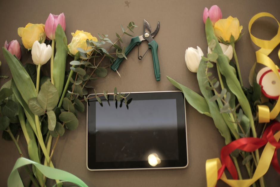 A flat lay arrangement of fresh flower bouquets featuring pink and yellow tulips with green leaves and eucalyptus sprigs, placed on a plain surface. Intertwined colorful ribbons in red, yellow, and gr