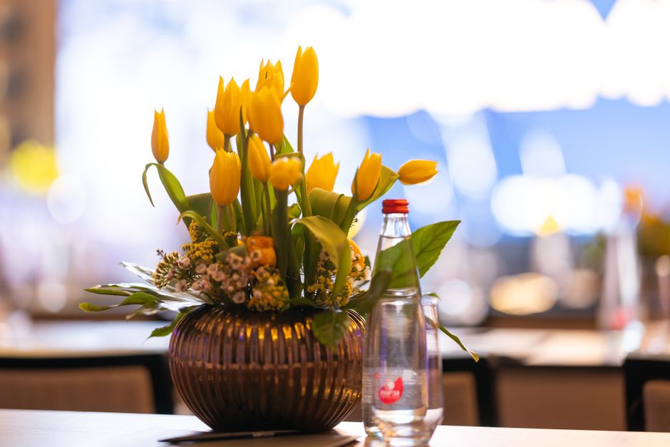 A floral arrangement consisting of bright yellow tulips with closed buds and open blooms, accompanied by small pink and purple filler flowers, is displayed in a round, ribbed amber glass vase on a tab