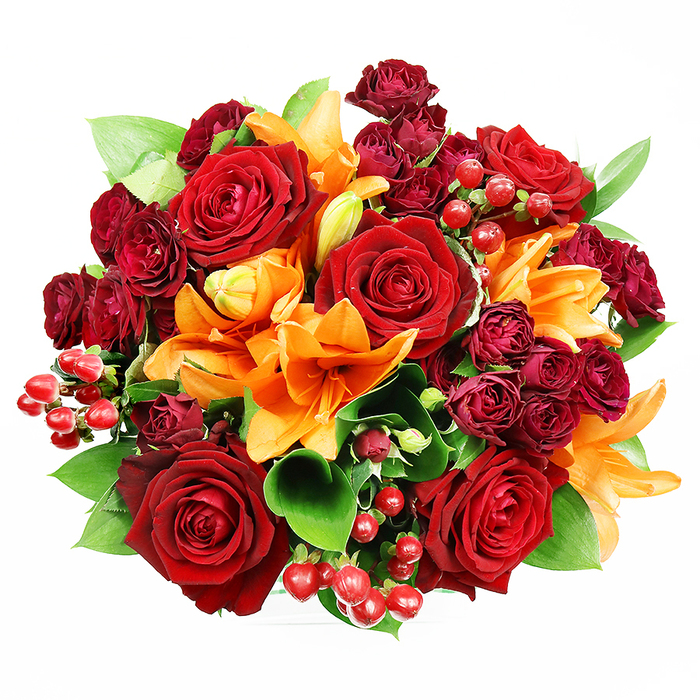 From a direct overhead viewpoint on a stark white surface, this opulent hand-tied bouquet presents itself as a jewel box of autumnal romance. Several large, fully opened deep red roses command attention with lush, velveteen petals curling outward to reveal complex, shadowed centres that seem to breathe warmth into the arrangement. Interspersed throughout are bright orange Asiatic lilies whose trumpet-shaped blooms and prominent stamens introduce dynamic lines and a zesty, almost citrus-spiced scent that lifts the deeper rose notes. Tucked close to the principal blooms, clusters of small burgundy spray roses add concentrated pockets of saturated colour and soft, textural contrast; their miniature heads create a plush, layered surface that reads like fine fabric against the smoother faces of the larger roses. Scattered like tiny rubies, glossy hypericum berries provide reflective highlights and a playful, festive rhythm across the bouquet's surface. Lush green foliage - the broad, glossy leaves of ruscus or salal - emerges between and beneath the flowers, framing the composition and amplifying the vibrant reds and oranges with fresh verdant tones. The studio lighting is gentle and diffuse, accentuating petal detail and the sheen of berries without producing harsh shadows, which gives the bouquet a polished, photographable quality ideal for gifting or editorial use. This arrangement conveys feelings of celebration, deep affection, and seasonal warmth; pictured here as a typical offering from a local Cubitt Town florist, it would sit beautifully on a mantel near Ferry Street or as the centrepiece for a small anniversary supper close to Canary Wharf, crafted to comfort the eye and the senses with every glance.